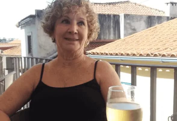 Uma senhora sorridente sentada, e sobre a mesa uma taça com champanhe